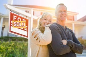 Attractive Middle-aged Couple In Front House and Sold Real Estate Sign.